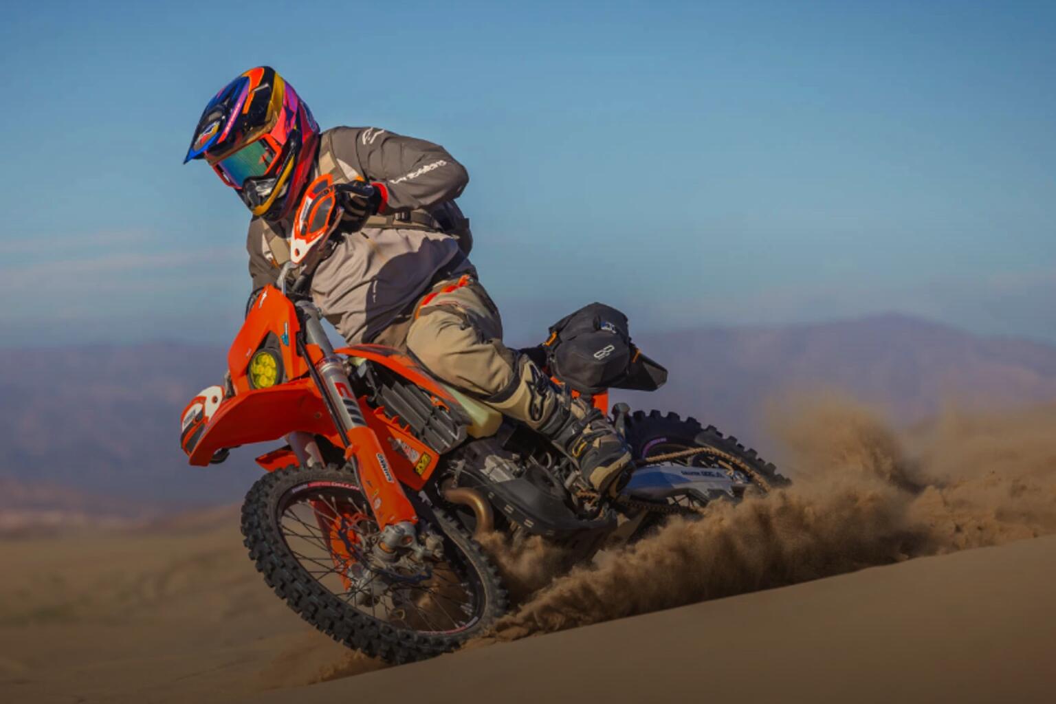 A rider on an orange dual sport carving a sandy desert turn, a wall of dust flying behind
