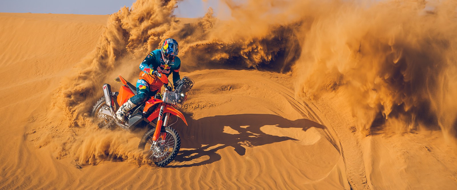 A KTM 450 Rally Replica rider carving a sand dune, spray flying