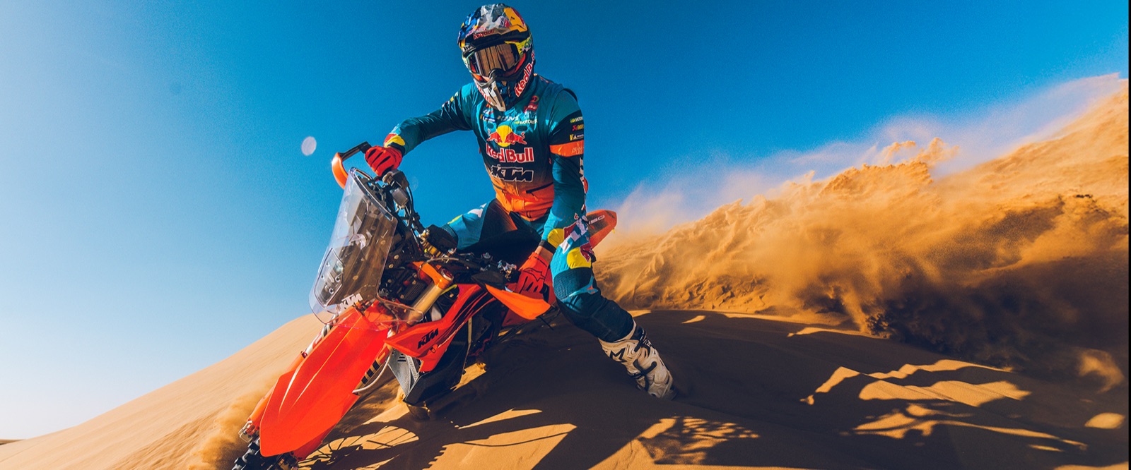 A low-angle shot of a KTM rider on a dune crest under a deep blue sky