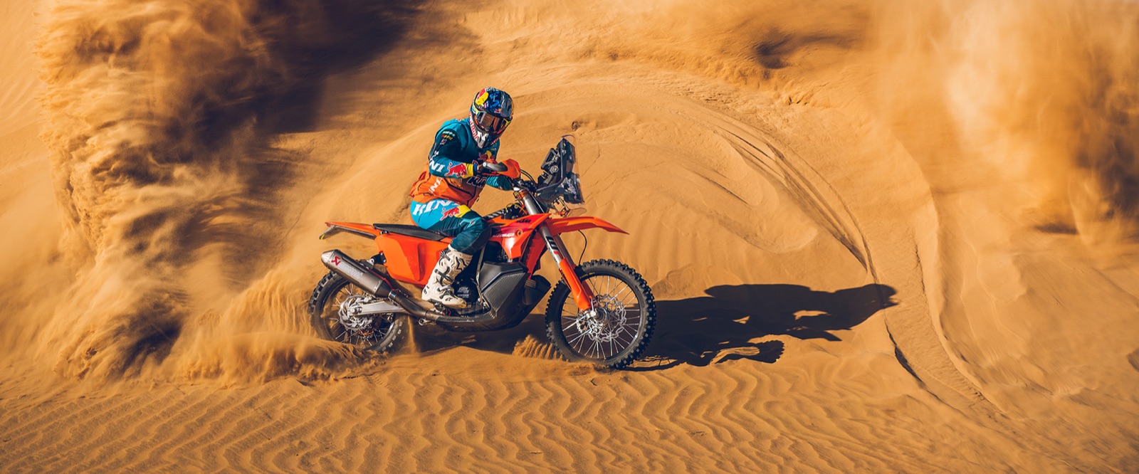 A KTM rider crests a sand dune with spray rising behind the bike