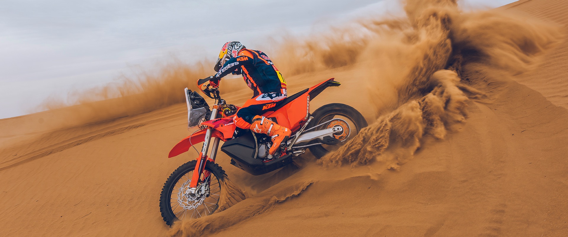 A KTM rider carves a dune and throws a wave of sand into the air