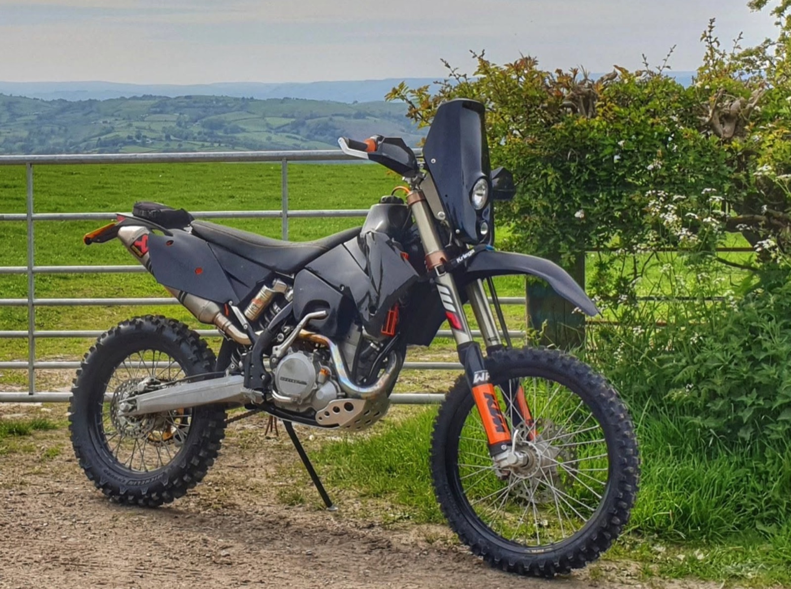 A grey KTM with a rally headlight screen at a farm gate, green hills behind