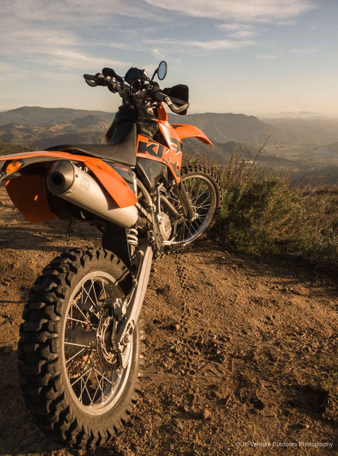 A KTM 525 EXC parked on a dirt ridge at sunset, hills behind