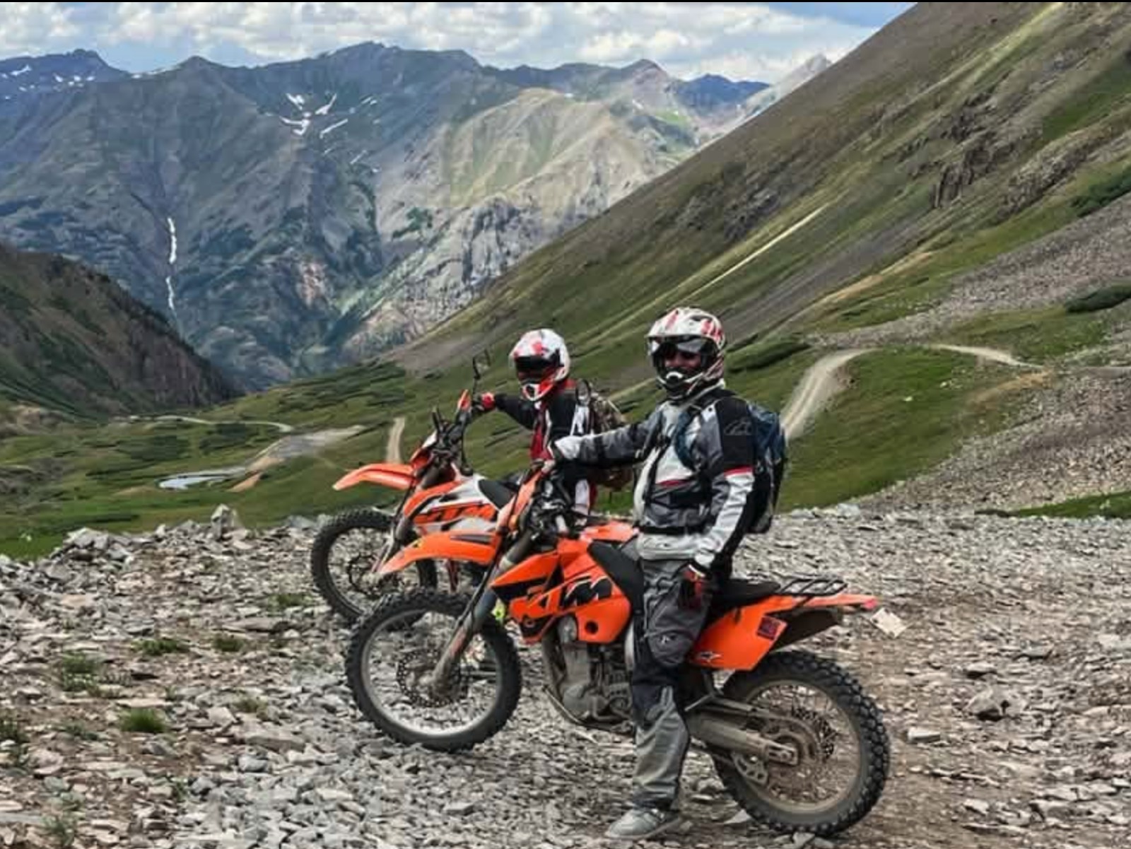 Two riders on orange KTMs stopped on a rocky mountain pass above a green valley