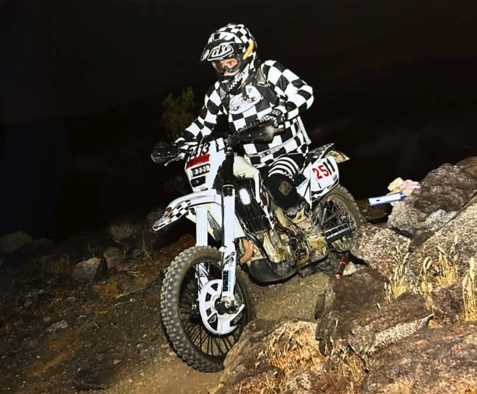 A rider in checkered gear on a white race-numbered bike climbing rocks at night