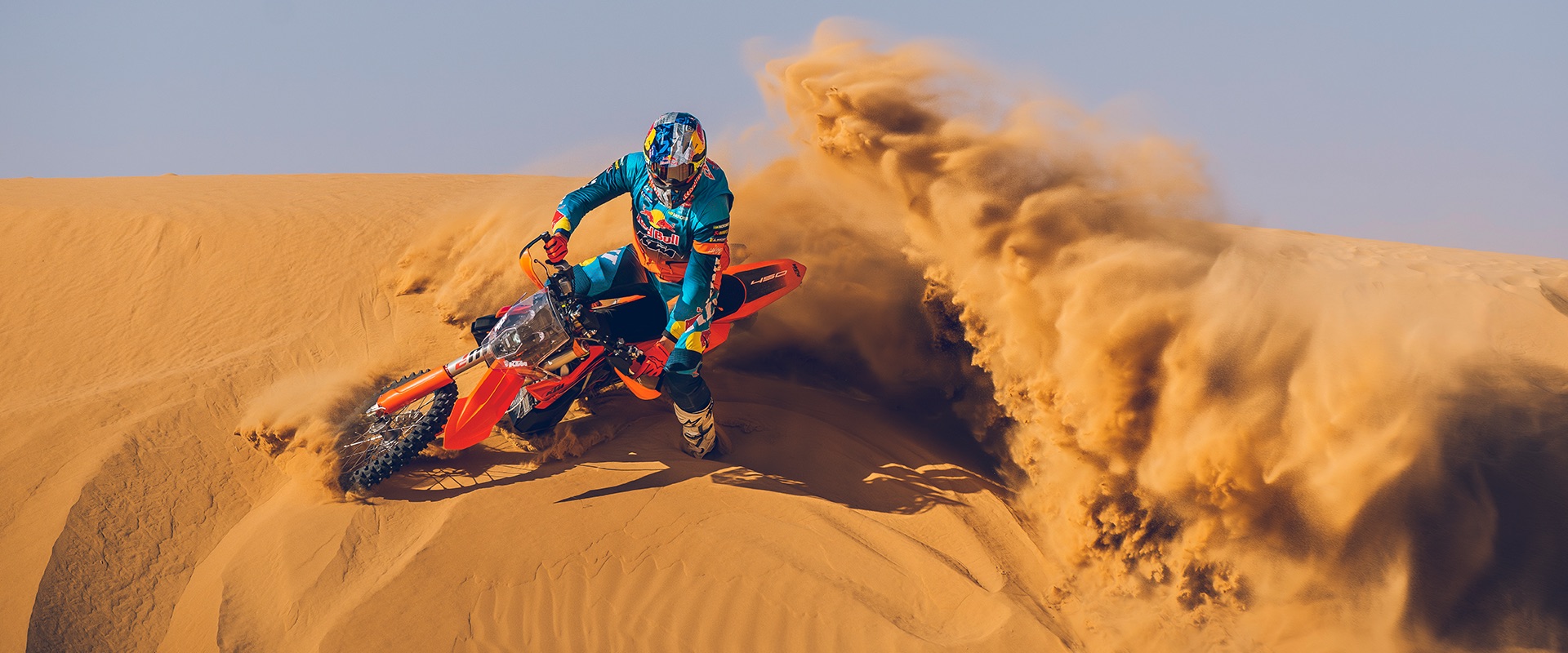 A KTM rider leans into a dune, throwing a wall of sand